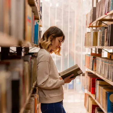Woman reading a book while standing between bookshelves in a library with natural light from large windows