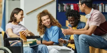 A group of four diverse students sitting and chatting together in a library or study area. One student is in a wheelchair, and others are holding books and notebooks. They are smiling and engaged in conversation.