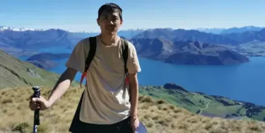 IDP IELTS test taker Liwei outdoors - A man with backpack and hiking pole standing on a grassy hilltop, overlooking a lake and snow-capped mountains under a clear blue sky
