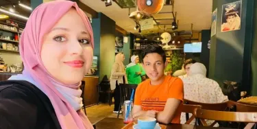 Group of people sitting in a café. In the foreground, two individuals, including Ahmed, are seated at a table with drinks - one wearing a pink headscarf, the other an orange shirt. Background includes more people, shelves with items, and posters on the walls