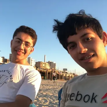 Two individuals, including Ahmed, sitting on a beach with buildings in the background. One wears a white t-shirt with the text 'SILENCE WILL NOT' and an illustration; the other wears a grey Reebok t-shirt