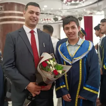 Two individuals standing side by side at a graduation ceremony. One wears a suit and holds a bouquet of flowers; Ahmed is in a graduation gown with blue and yellow accents. Background includes other people in similar graduation attire