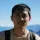 IDP IELTS test taker Liwei outdoors - A man wearing a beige shirt and a backpack, standing outdoors with snow-capped mountains in the background under a clear blue sky